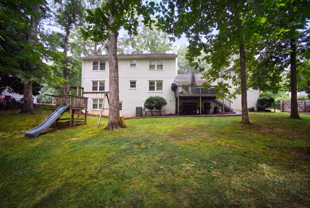 Rear exterior with elevated deck, covered patio, playset, and expansive tree-shaded backyard